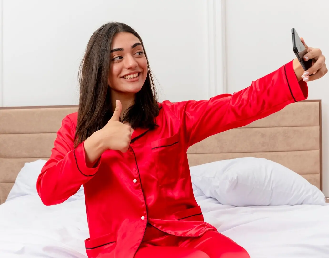 Junge schöne Frau im roten Pyjama sitzt auf dem Bett, benutzt ein Smartphone, macht ein Selfie, lächelt und zeigt den Daumen hoch, im Schlafzimmerinterieur vor hellem Hintergrund.